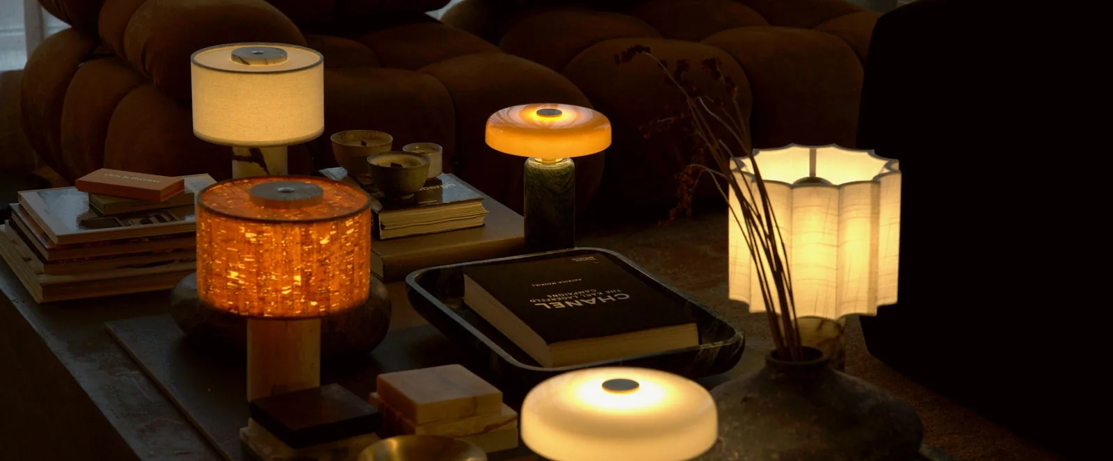 Five Amberden lamps lit on a low table, a velvet sofa behind.